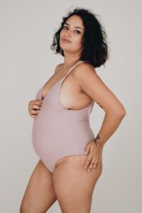Model wearing light pink one piece holding her stomach.